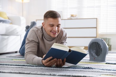 Photo of Young man reading book near electric heater at home