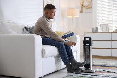 Photo of Young man reading book near electric heater at home