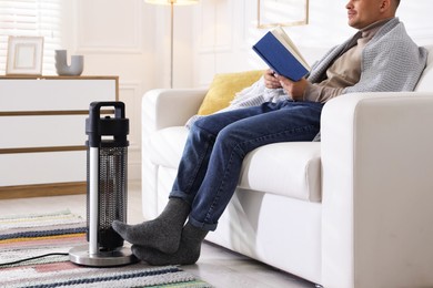 Photo of Young man reading book near electric heater at home, closeup
