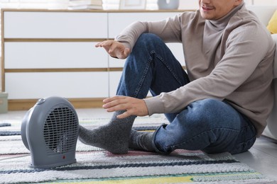 Photo of Young man near electric heater on floor at home, closeup