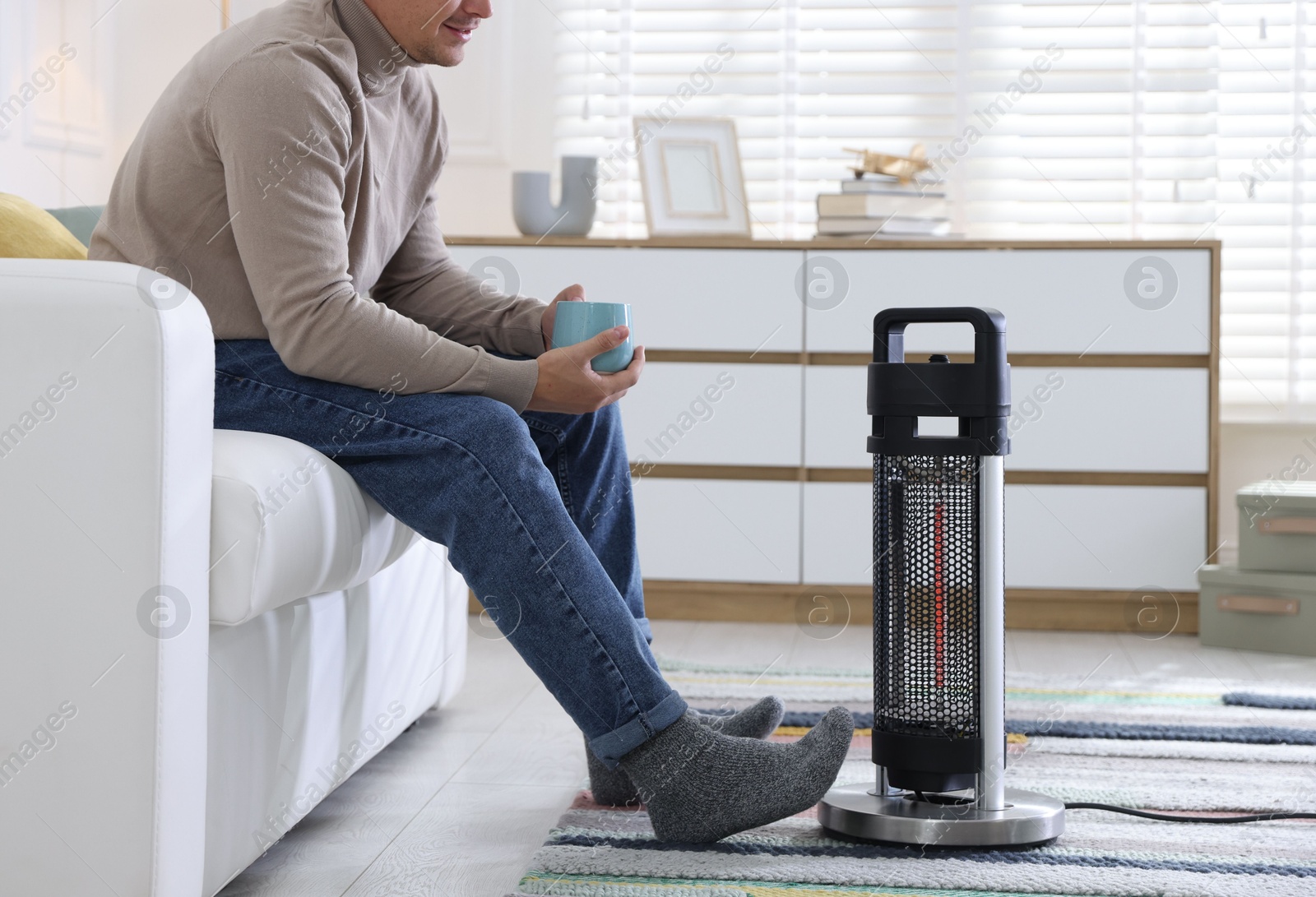 Photo of Young man with cup of drink near electric heater at home, closeup