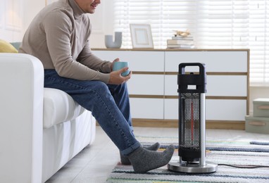 Photo of Young man with cup of drink near electric heater at home, closeup