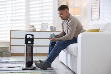 Photo of Young man with cup of drink near electric heater at home