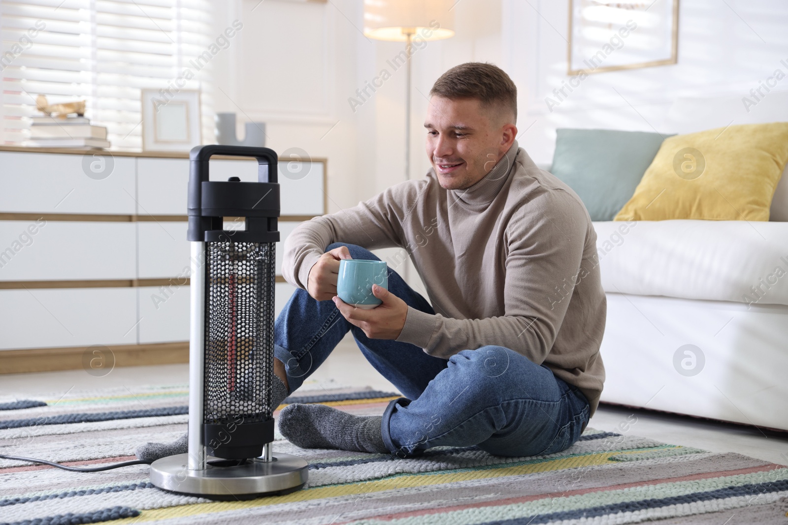 Photo of Young man with cup of drink near electric heater at home