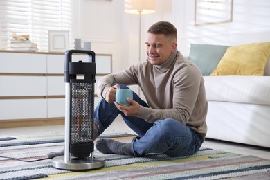 Photo of Young man with cup of drink near electric heater at home