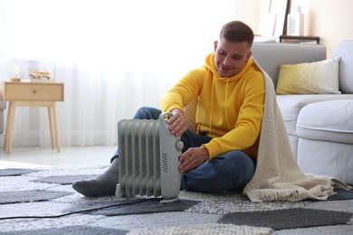 Photo of Young man with blanket near electric heater on floor at home