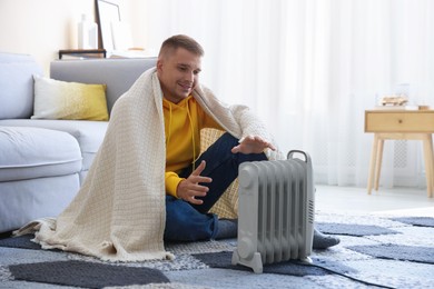 Photo of Young man with blanket near electric heater on floor at home