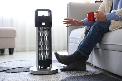 Photo of Young man with cup of drink near electric heater at home, closeup