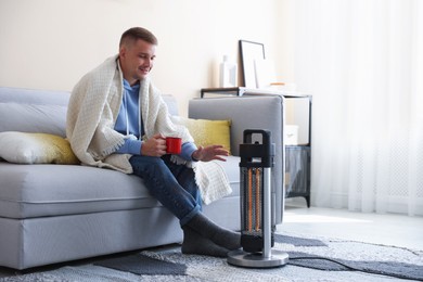 Photo of Young man with cup of drink near electric heater at home