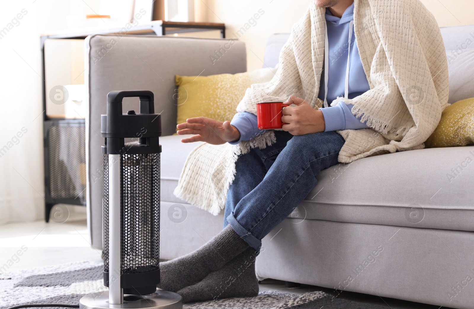 Photo of Young man with cup of drink near electric heater at home, closeup