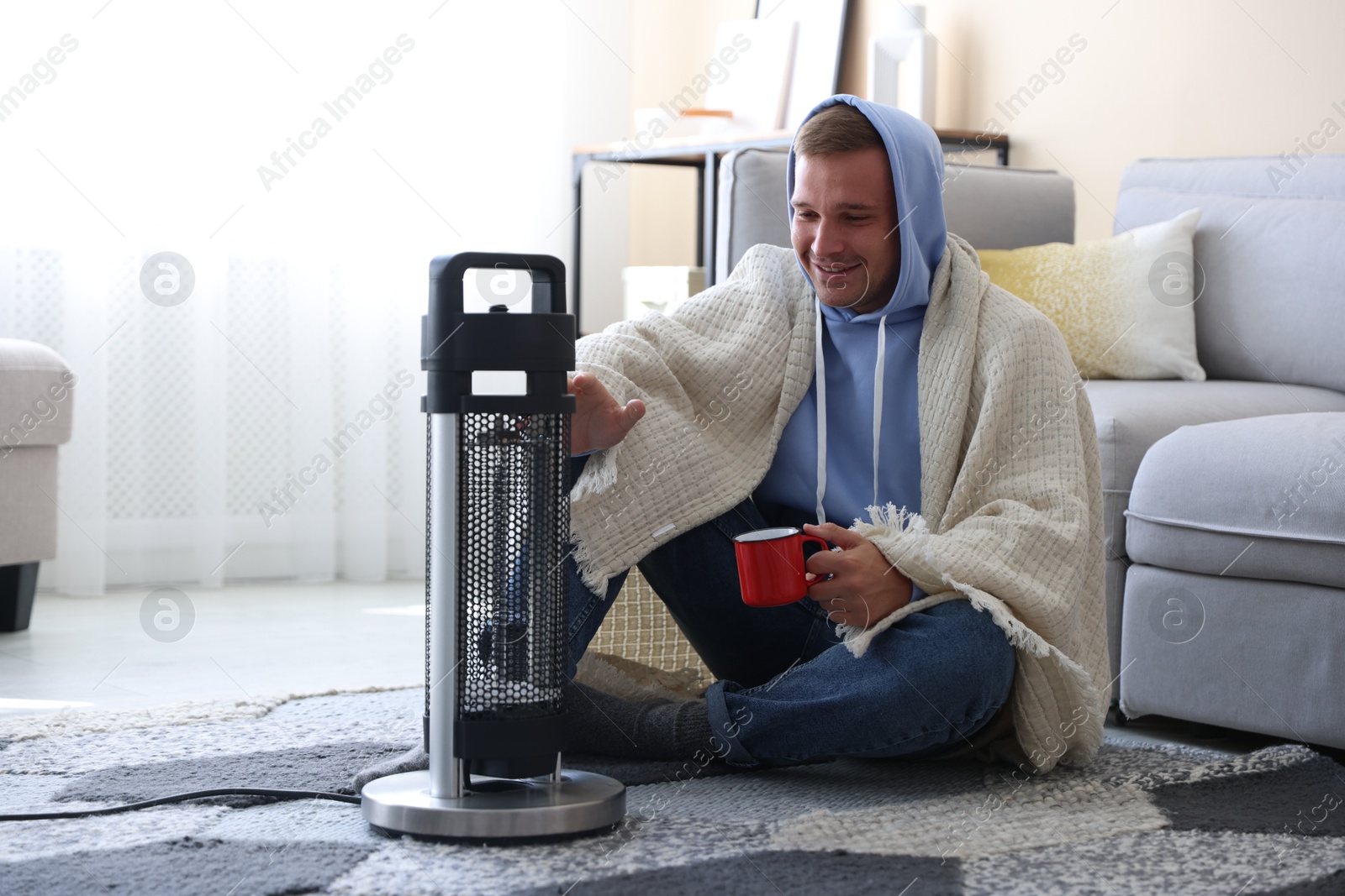 Photo of Young man with cup of drink near electric heater at home