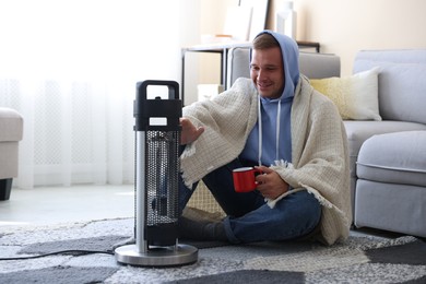 Photo of Young man with cup of drink near electric heater at home