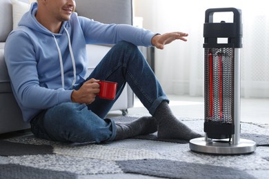 Photo of Young man with cup of drink near electric heater at home, closeup
