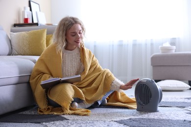Photo of Young woman reading book near electric heater at home