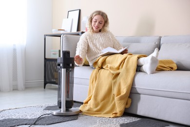 Photo of Young woman reading book near electric heater at home