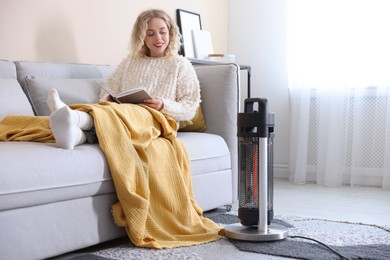 Photo of Young woman reading book near electric heater at home