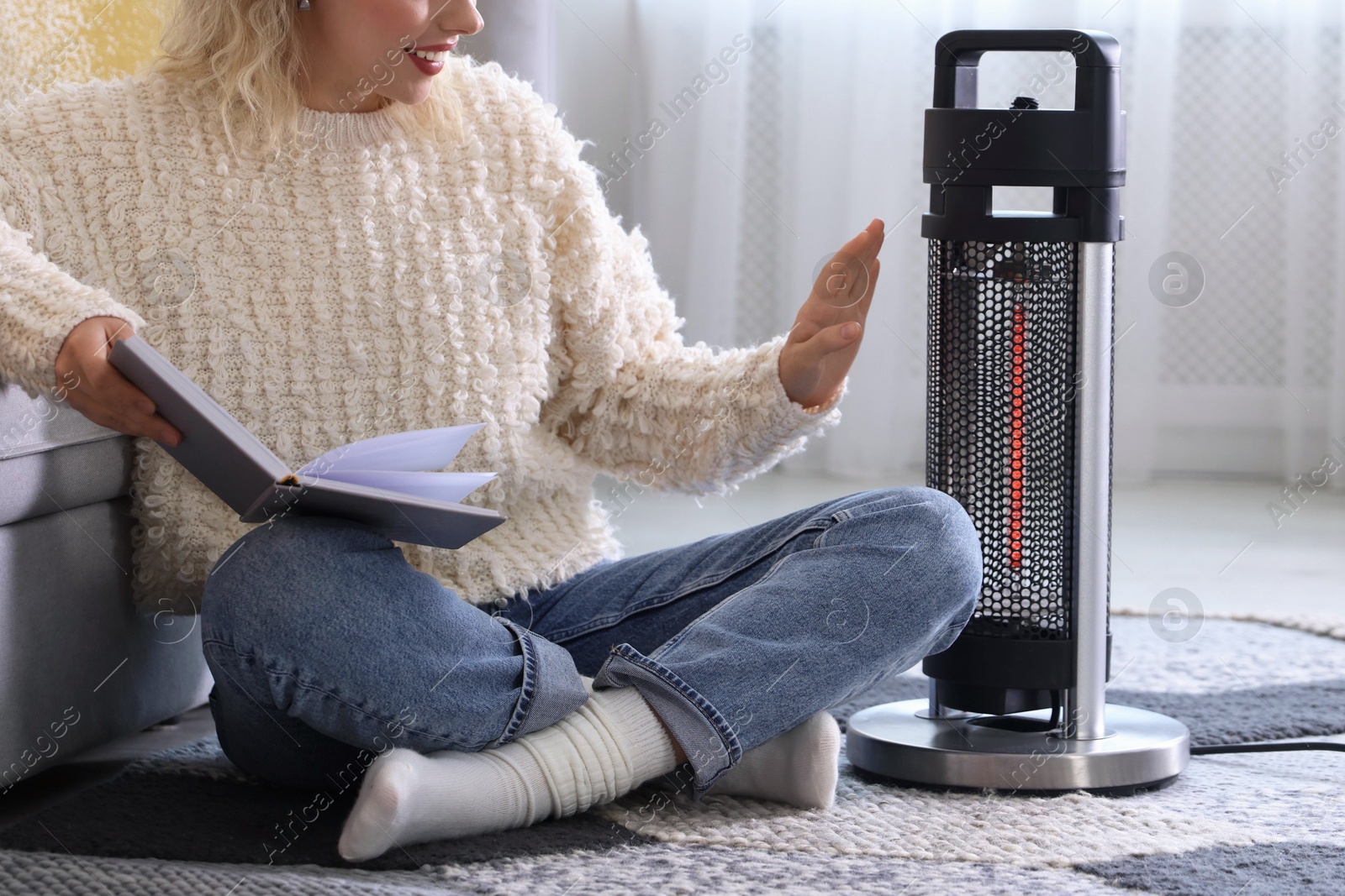 Photo of Young woman reading book near electric heater at home, closeup