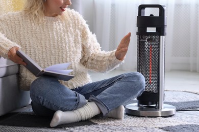 Photo of Young woman reading book near electric heater at home, closeup