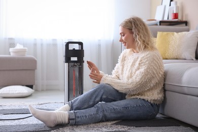 Photo of Young woman near electric heater on floor at home