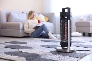 Photo of Young woman with cup of drink and book near electric heater on floor at home, selective focus