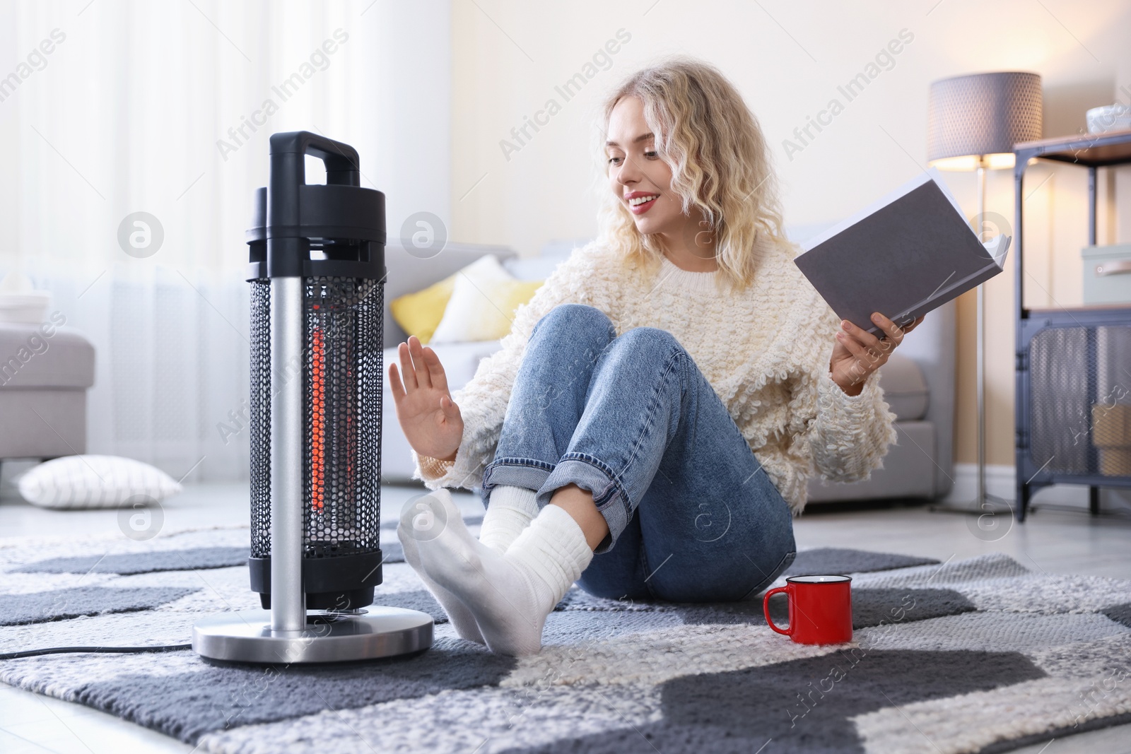 Photo of Young woman with cup of drink and book near electric heater on floor at home