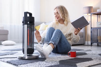 Photo of Young woman with cup of drink and book near electric heater on floor at home