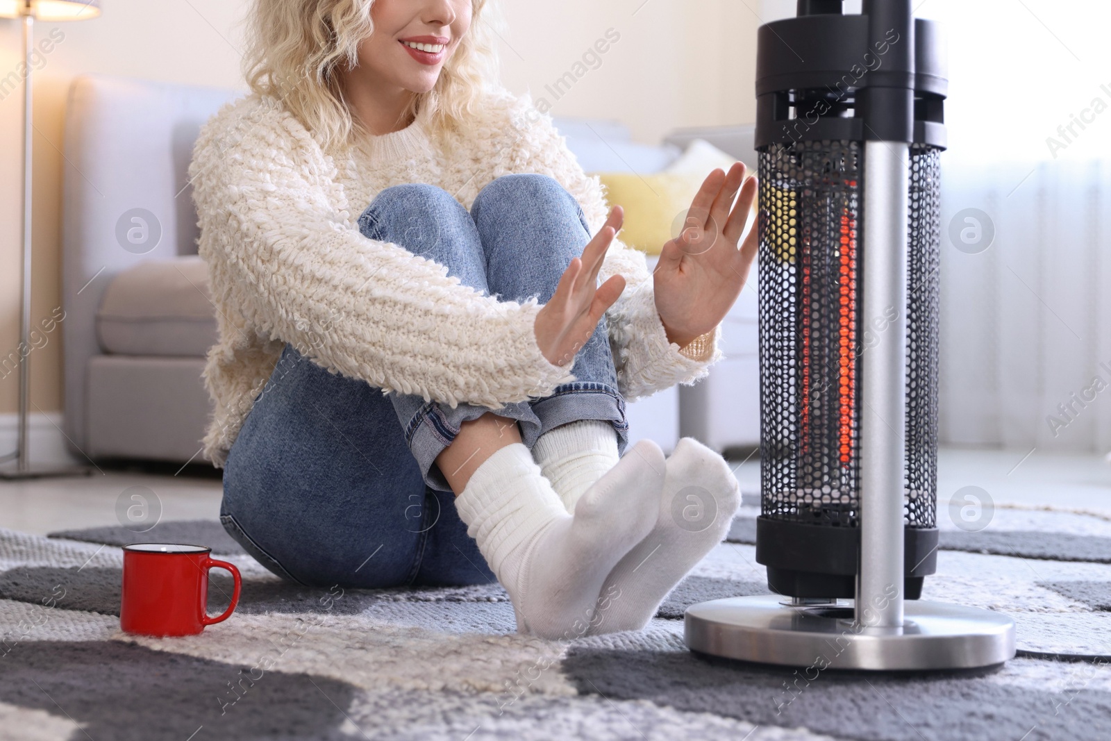 Photo of Young woman with cup of drink near electric heater on floor at home, closeup
