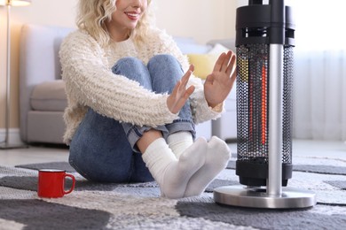 Photo of Young woman with cup of drink near electric heater on floor at home, closeup