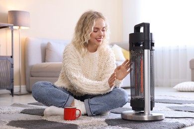 Photo of Young woman with cup of drink near electric heater on floor at home