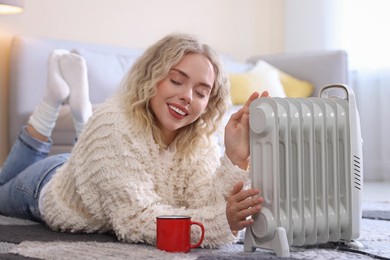 Photo of Young woman with cup of drink near electric heater on floor at home