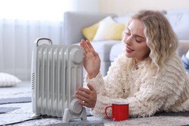 Photo of Young woman with cup of drink near electric heater on floor at home