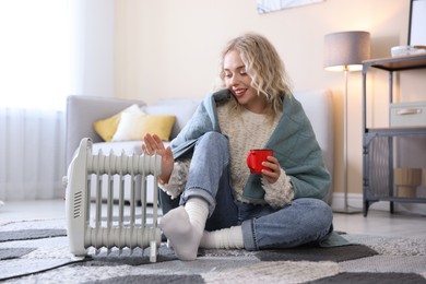 Photo of Young woman with cup of drink near electric heater on floor at home