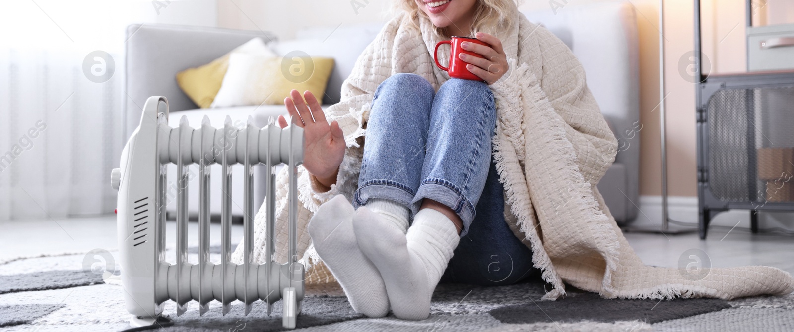 Photo of Young woman with cup of drink near electric heater on floor at home, closeup