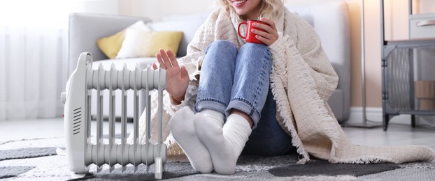 Photo of Young woman with cup of drink near electric heater on floor at home, closeup