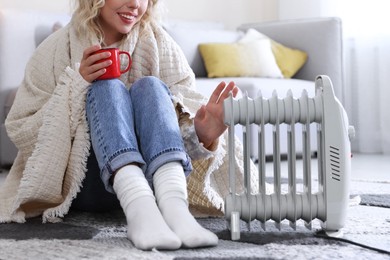 Photo of Young woman with cup of drink near electric heater on floor at home, closeup