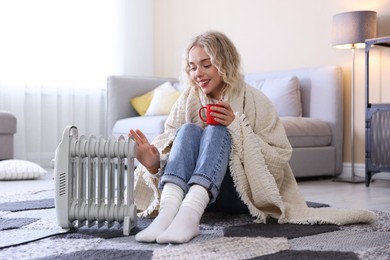 Photo of Young woman with cup of drink near electric heater on floor at home