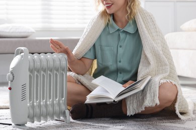 Photo of Young woman with book near electric heater on floor at home, closeup