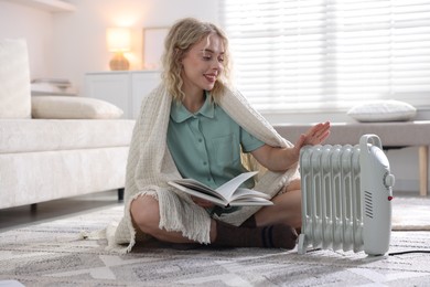 Photo of Young woman with book near electric heater on floor at home