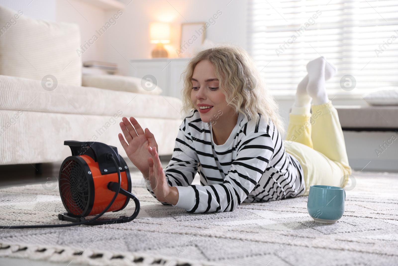 Photo of Young woman with cup of drink near electric heater at home