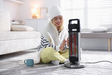 Photo of Young woman with cup of drink near electric heater at home