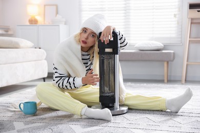 Photo of Young woman with cup of drink near electric heater at home
