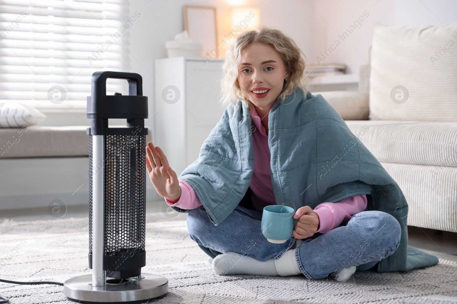Photo of Young woman with cup of drink near electric heater at home