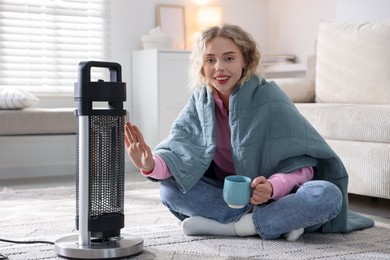 Photo of Young woman with cup of drink near electric heater at home