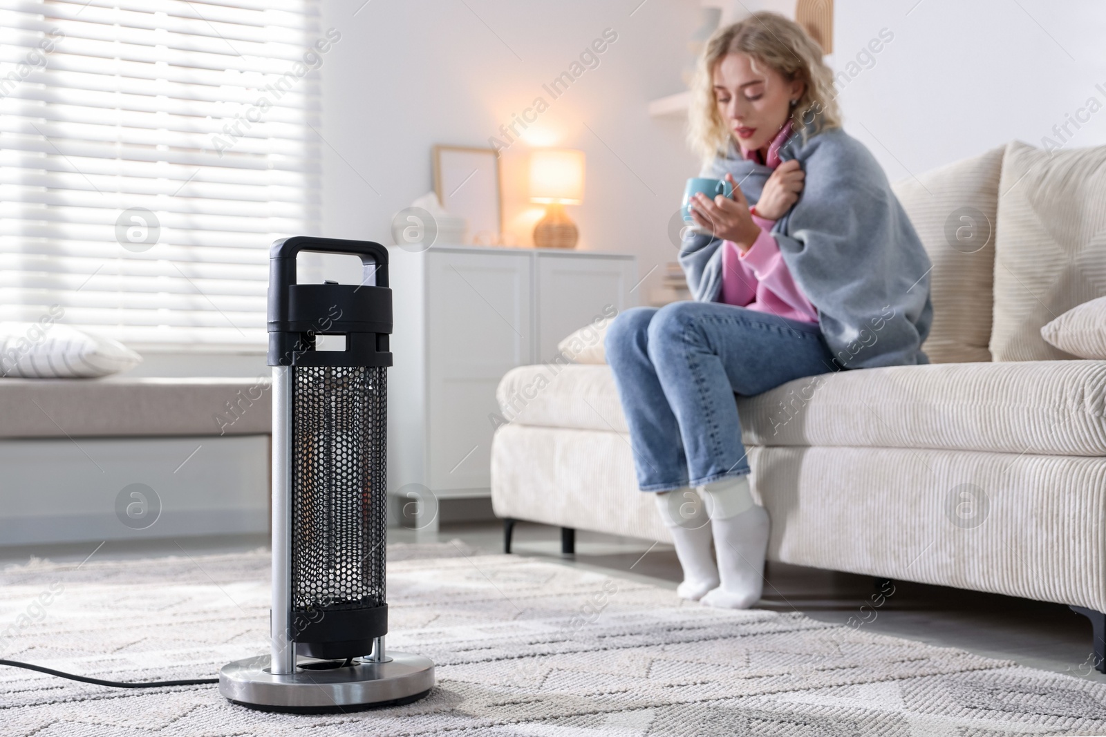 Photo of Young woman with cup of drink near electric heater at home, selective focus