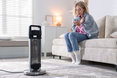 Photo of Young woman with cup of drink near electric heater at home, selective focus