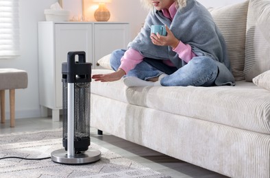 Photo of Young woman with cup of drink near electric heater at home, closeup