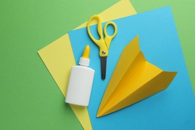 Photo of Glue in bottle, scissors, color paper and handmade plane on green background, flat lay. Space for text