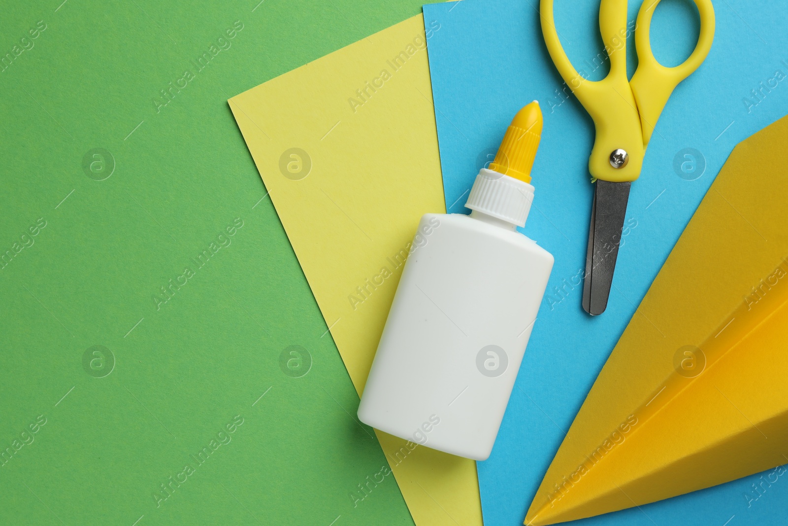 Photo of Glue in bottle, scissors, color paper and handmade plane on green background, flat lay. Space for text