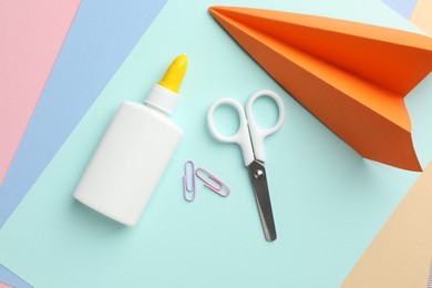 Photo of Glue in bottle, scissors, handmade plane and paper clips on color paper, flat lay