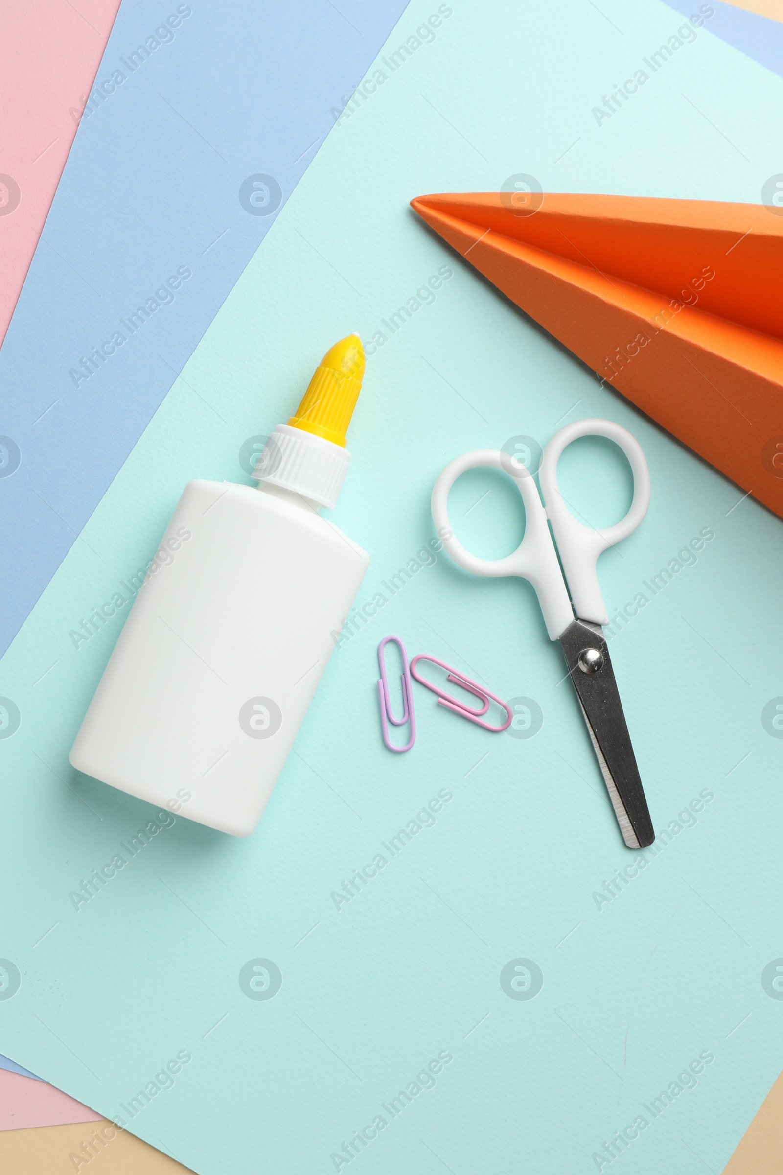 Photo of Glue in bottle, scissors, handmade plane and paper clips on color paper, flat lay